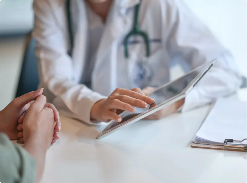 A doctor in a white coat points to something on a tablet, showing a patient.